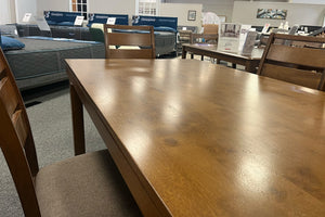 Vista cercana de mesa de comedor rectangular en acabado madera nogal con superficie amplia y veta visible. Rodeada de sillas a juego con asientos tapizados, el conjunto se exhibe en una tienda de muebles y destaca por su diseño clásico y funcional.