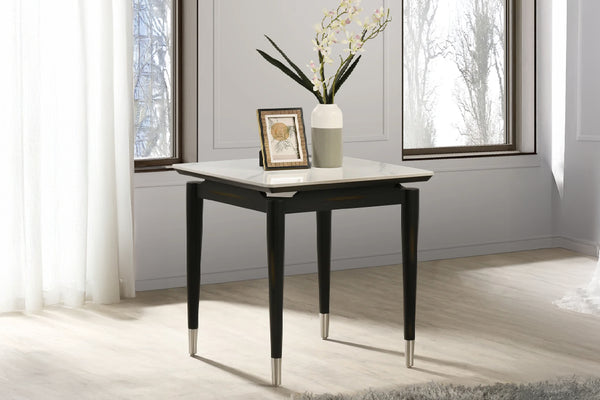 Una mesa auxiliar con tope cuadrado de imitación de mármol blanco y patas negras y cónicas, cada una rematada con una virola de plata o cromo. La mesa está colocada frente a grandes ventanas con cortinas blancas transparentes. Sobre la mesa hay un arreglo decorativo que incluye un jarrón con orquídeas blancas y amarillas y un pequeño marco de fotos.