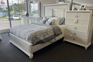Juego de dormitorio en acabado blanco con cama de tamaño queen, cabecera tapizada y detalles decorativos brillantes. Incluye cómoda con espejo y gavetero alto a juego, complementados con ropa de cama en tonos grises y diseño elegante.