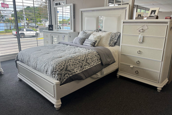 Juego de dormitorio en acabado blanco con cama de tamaño queen, cabecera tapizada y detalles decorativos brillantes. Incluye cómoda con espejo y gavetero alto a juego, complementados con ropa de cama en tonos grises y diseño elegante.