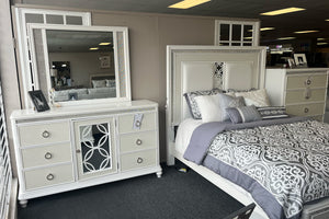 Juego de dormitorio elegante en acabado blanco exhibido en tienda, con cama de cabecera tapizada con paneles y detalles decorativos. Incluye gavetero con seis gavetas y espejo rectangular a juego, con tiradores metálicos circulares y detalles brillantes. La cama está vestida en tonos gris y blanco con patrón ornamental y cojines decorativos.