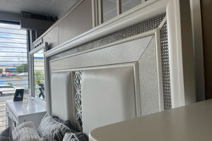 Detalle de cabecera de cama en acabado blanco con paneles acolchonados y marco decorativo con incrustaciones brillantes. Se aprecian cojines en tonos gris y blanco, con espejo y mobiliario a juego reflejados en un ambiente de tienda.