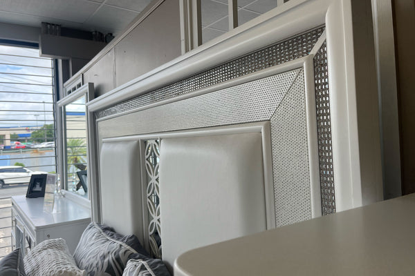 Detalle de cabecera de cama en acabado blanco con paneles acolchonados y marco decorativo con incrustaciones brillantes. Se aprecian cojines en tonos gris y blanco, con espejo y mobiliario a juego reflejados en un ambiente de tienda.