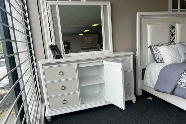 Gavetero con espejo a juego en acabado blanco con detalles decorativos brillantes, exhibido en tienda. Incluye tres gavetas a cada lado con tiradores circulares metálicos y un compartimento central con puerta abatible abierta que muestra espacio de almacenamiento interno.
