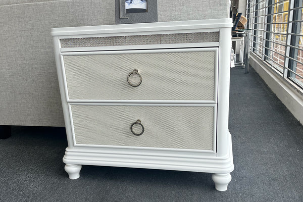 Mesa de noche de dos gavetas en acabado blanco con frentes texturizados y tiradores metálicos en forma de aro. Su diseño clásico incluye molduras decorativas y patas torneadas, aportando elegancia y funcionalidad al dormitorio.