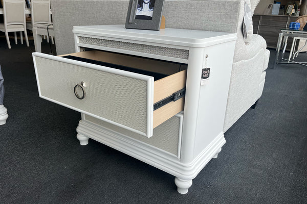 Mesa de noche de dos gavetas en acabado blanco con frentes texturizados y tiradores metálicos tipo aro. La gaveta superior aparece abierta, mostrando su capacidad de almacenamiento y destacando el diseño clásico con patas torneadas.