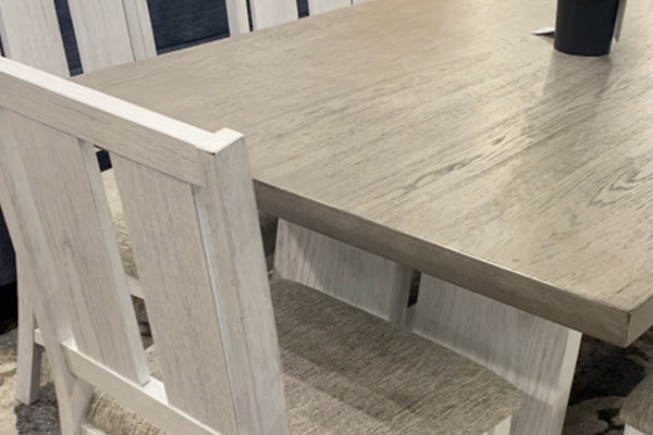 Primer plano de mesa de comedor con acabado de madera en tonos gris claro y veta visible, junto a una silla a juego con respaldo de listones y asiento tapizado en tela beige. El diseño combina estilo rústico y funcionalidad para espacios acogedores.