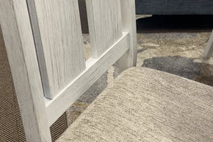 Primer plano de silla de comedor con acabado blanco envejecido, respaldo de listones verticales y asiento tapizado en tela beige texturizada. El diseño combina detalles rústicos y tonos suaves para un estilo acogedor y versátil.