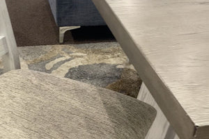 Detalle de mesa de comedor con acabado de madera en tonos gris claro y textura de veta visible, junto a asiento tapizado en tela beige. La combinación de materiales y colores suaves aporta un estilo rústico moderno y acogedor.