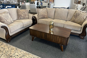 Loveseat sofá marrón con cojines estampados y una mesa de centro de madera en un ambiente de sala de exposición.