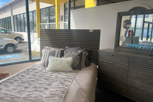 Juego de dormitorio moderno en acabado madera gris exhibido en tienda, con cama de cabecera con paneles horizontales, ropa de cama en tonos beige y cojines decorativos. Incluye gavetero con múltiples gavetas y espejo a juego, ubicado junto a ventanales con vista exterior y luz natural.