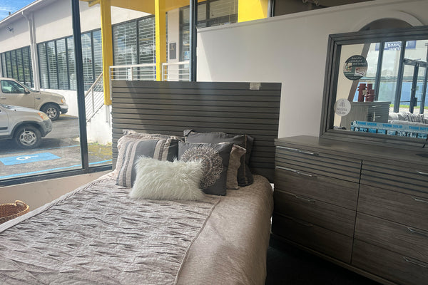 Juego de dormitorio moderno en acabado madera gris exhibido en tienda, con cama de cabecera con paneles horizontales, ropa de cama en tonos beige y cojines decorativos. Incluye gavetero con múltiples gavetas y espejo a juego, ubicado junto a ventanales con vista exterior y luz natural.