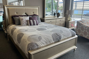 Juego de dormitorio contemporáneo en acabado beige exhibido en tienda, con cama de cabecera tapizada con paneles acolchonados. La cama está vestida con edredón blanco con círculos grises y cojines decorativos en tonos neutros y morado. Incluye mesa de noche y gavetero con tiradores metálicos, junto a ventanas con luz natural.