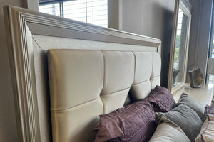Detalle de cabecera de cama tapizada en tono beige con paneles acolchonados geométricos y marco en acabado plateado. Incluye cojines decorativos en tonos morado y gris, con fondo de ventana y espejo en un entorno de tienda.