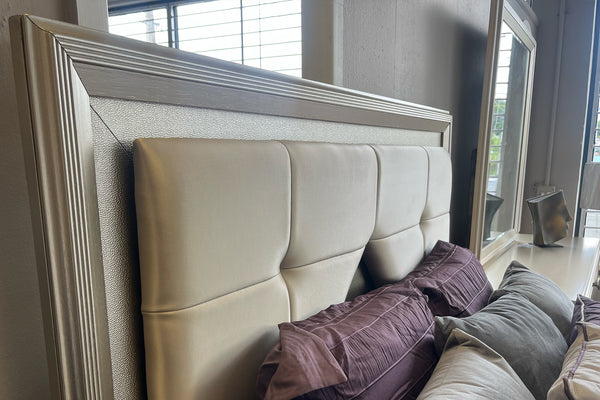 Detalle de cabecera de cama tapizada en tono beige con paneles acolchonados geométricos y marco en acabado plateado. Incluye cojines decorativos en tonos morado y gris, con fondo de ventana y espejo en un entorno de tienda.