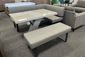 Juego de comedor tipo banco esquinero en color gris con mesa rectangular de acabado brillante, banco tapizado y base geométrica metálica. Exhibido en una sala de muebles contemporáneos con sofás y dormitorios al fondo.