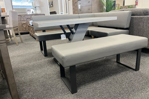 Conjunto de comedor moderno con banco esquinero en forma de L, banco auxiliar tapizado y mesa rectangular gris de acabado brillante. La mesa cuenta con base geométrica metálica y se exhibe en una tienda de muebles contemporáneos.
