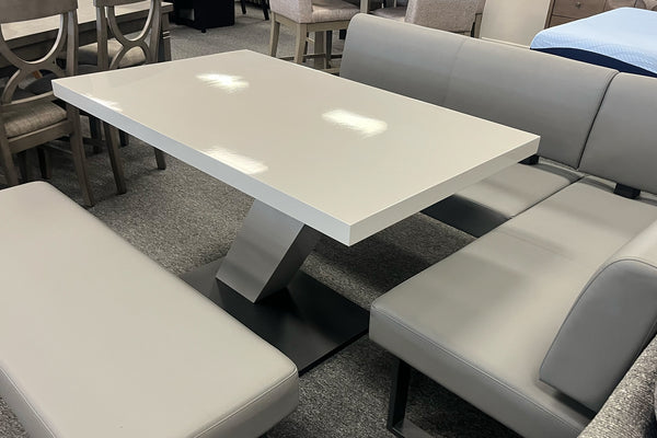 Juego de comedor contemporáneo con mesa rectangular blanca de acabado brillante, base central geométrica en tono gris y asientos tapizados grises. Incluye banco auxiliar y banco esquinero en forma de L para maximizar el espacio.