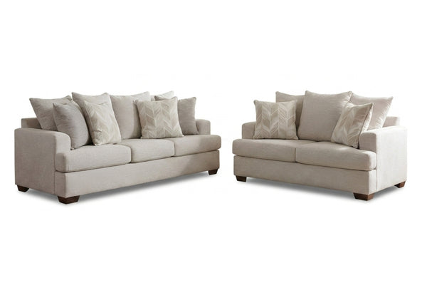 Juego de sala compuesto por sofá y loveseat tapizados en tela color beige claro, con cojines decorativos a juego y patas de madera oscura. Presentado sobre fondo blanco para resaltar el diseño contemporáneo y las líneas limpias del conjunto.