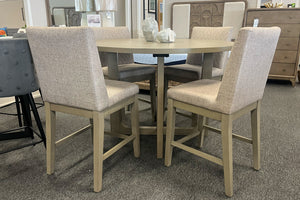 Juego de comedor de altura de mostrador con mesa redonda en acabado gris claro y cuatro sillas tapizadas en tela color beige. El conjunto presenta una base central geométrica y un diseño contemporáneo ideal para espacios modernos y acogedores.