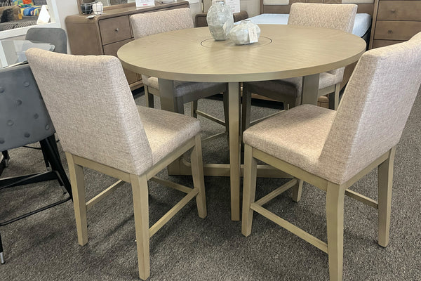 Juego de comedor de altura de mostrador con mesa redonda en acabado gris claro y cuatro sillas tapizadas en tela color beige. Presenta una base central geométrica y diseño contemporáneo, exhibido en una sala de muebles con estilo moderno y tonos neutros.