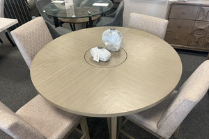 Vista superior de una mesa redonda de comedor en acabado gris claro con detalle central giratorio tipo lazy Susan. Rodeada por cuatro sillas tapizadas en tela color beige con respaldo acolchado, exhibida en una sala de muebles contemporánea.