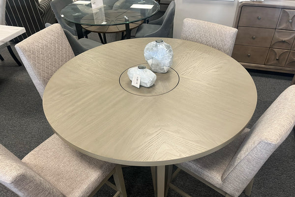 Vista superior de una mesa redonda de comedor en acabado gris claro con detalle central giratorio tipo lazy Susan. Rodeada por cuatro sillas tapizadas en tela color beige con respaldo acolchado, exhibida en una sala de muebles contemporánea.