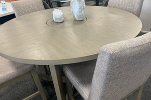 Primer plano de una mesa redonda de comedor en acabado gris claro con centro giratorio integrado tipo lazy Susan. Rodeada por sillas tapizadas en tela beige con respaldo acolchado, destacando la veta de la madera y el diseño contemporáneo.
