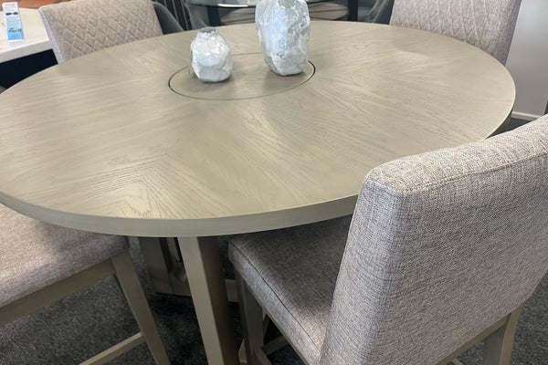 Primer plano de una mesa redonda de comedor en acabado gris claro con centro giratorio integrado tipo lazy Susan. Rodeada por sillas tapizadas en tela beige con respaldo acolchado, destacando la veta de la madera y el diseño contemporáneo.