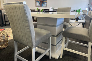 Juego de comedor tipo pub en acabado blanco brillante con detalles decorativos en brillo plateado. Incluye mesa cuadrada de pedestal central y sillas altas tapizadas en terciopelo gris con respaldo acolchado vertical, exhibido en una sala de muebles.