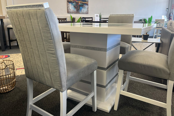 Juego de comedor tipo pub en acabado blanco brillante con detalles decorativos en brillo plateado. Incluye mesa cuadrada de pedestal central y sillas altas tapizadas en terciopelo gris con respaldo acolchado vertical, exhibido en una sala de muebles.