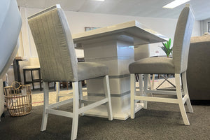 Vista lateral de un juego de comedor tipo pub con mesa cuadrada en acabado blanco brillante y pedestal central con detalles plateados. Incluye sillas altas tapizadas en gris claro con respaldo acolchado vertical y estructura blanca.