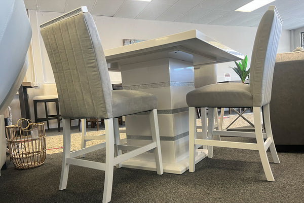 Vista lateral de un juego de comedor tipo pub con mesa cuadrada en acabado blanco brillante y pedestal central con detalles plateados. Incluye sillas altas tapizadas en gris claro con respaldo acolchado vertical y estructura blanca.