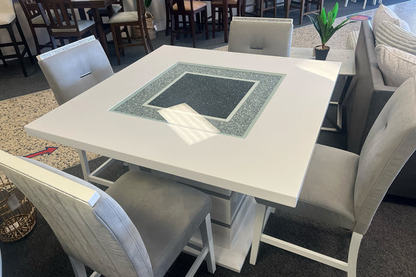 Vista superior de un juego de comedor tipo pub con mesa cuadrada en acabado blanco brillante y panel central decorativo con cristal y detalles brillantes. Incluye cuatro sillas altas tapizadas en gris claro, exhibidas en una sala de muebles contemporánea.