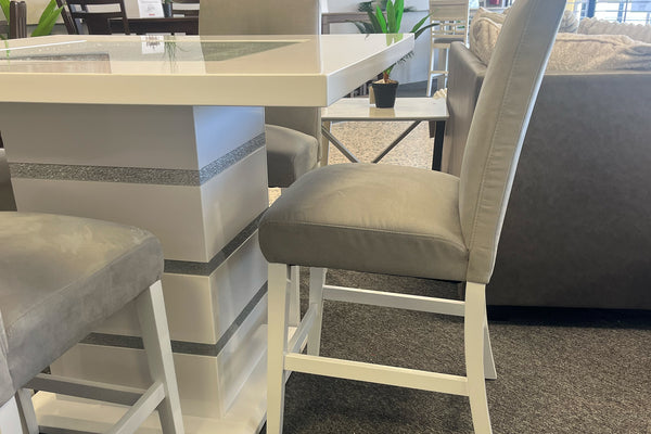 Primer plano de una silla alta tapizada en terciopelo gris claro junto a una mesa tipo pub en acabado blanco brillante. Se aprecia el pedestal central con detalles decorativos plateados y la estructura blanca del conjunto de comedor contemporáneo.