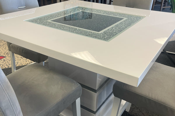 Primer plano de una mesa tipo pub en acabado blanco brillante con panel central de cristal decorativo y detalles brillantes. La imagen resalta la superficie de alto brillo, el pedestal geométrico y los asientos tapizados en gris claro.