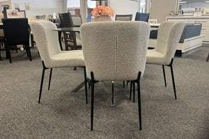 Juego de comedor con mesa redonda de vidrio y cuatro sillas tapizadas en tela tipo bouclé color crema. Las sillas cuentan con respaldo curvo y patas metálicas negras, exhibidas en una sala de exhibición de muebles.