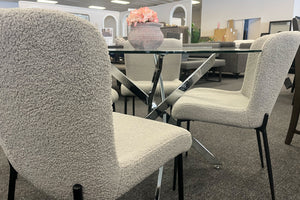 Detalle de un juego de comedor con mesa redonda de vidrio y base metálica cromada en forma de cruz. Rodeada por sillas tapizadas en tela bouclé color crema con patas negras, exhibidas en una tienda de muebles.