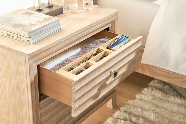 Mesita de noche de madera con una gaveta abierta que contiene joyas, libros encima y un teléfono.