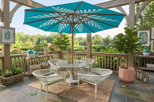 Conjunto de comedor para exterior en terraza cubierta, con mesa redonda, cuatro sillas blancas de cuerda tejida y sombrilla turquesa con diseño de hojas. Rodeado de plantas decorativas y vistas a un jardín verde.