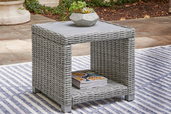 Outdoor end table with a gray woven frame and glass top in a patio setting. 