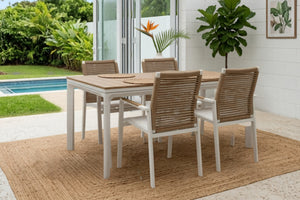 Juego de comedor para exterior con mesa rectangular en tonos claros y cuatro sillas con respaldo tejido. Ubicado en una terraza cubierta junto a una piscina, rodeado de plantas tropicales y abundante luz natural.