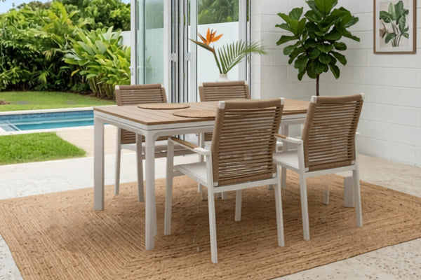 Juego de comedor para exterior con mesa rectangular en tonos claros y cuatro sillas con respaldo tejido. Ubicado en una terraza cubierta junto a una piscina, rodeado de plantas tropicales y abundante luz natural.