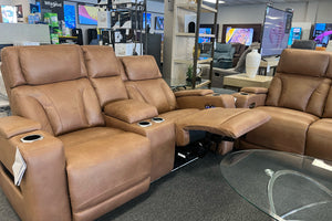 Sofá reclinable modular en color café claro con asientos acolchados, portavasos integrados y reposapiés extendido, exhibido junto a sillones reclinables en una sala de muebles.