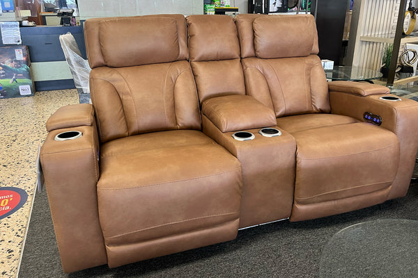 Loveseat reclinable en color café claro con respaldo acolchado, consola central con portavasos y controles laterales iluminados, exhibido en una tienda de muebles.