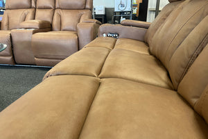 Primer plano de sofá reclinable en color café claro con asientos acolchados amplios, respaldo con costuras verticales y controles integrados en el reposabrazos, exhibido en sala de muebles.