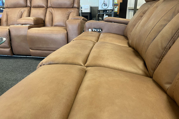 Primer plano de sofá reclinable en color café claro con asientos acolchados amplios, respaldo con costuras verticales y controles integrados en el reposabrazos, exhibido en sala de muebles.