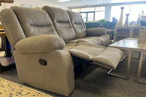 Sofá reclinable en color gris claro con respaldo acolchado, brazos amplios y reposapiés extendido, exhibido junto a una mesa de centro con acabado de madera clara en una tienda de muebles.