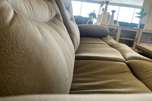 Primer plano lateral de sofá reclinable en color gris claro con tapizado texturizado, asientos acolchados y brazos amplios, exhibido en una tienda de muebles.