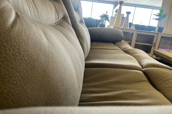Primer plano lateral de sofá reclinable en color gris claro con tapizado texturizado, asientos acolchados y brazos amplios, exhibido en una tienda de muebles.